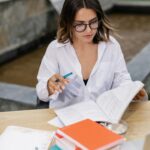 A young woman with eyeglasses studying outdoors with books and a laptop, portraying a studious and focused atmosphere.